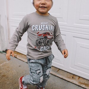 May include: A young child wearing a grey long-sleeve shirt with a red monster truck graphic and the text "Heart Crushing Crew". The child is also wearing a white beanie, distressed denim jeans, and red high-top sneakers.
