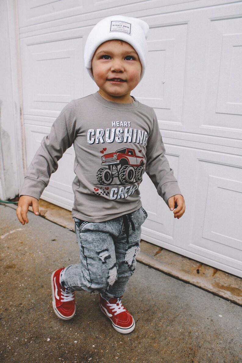 May include: A young child wearing a grey long-sleeve shirt with a red monster truck graphic and the text "Heart Crushing Crew". The child is also wearing a white beanie, distressed denim jeans, and red high-top sneakers.