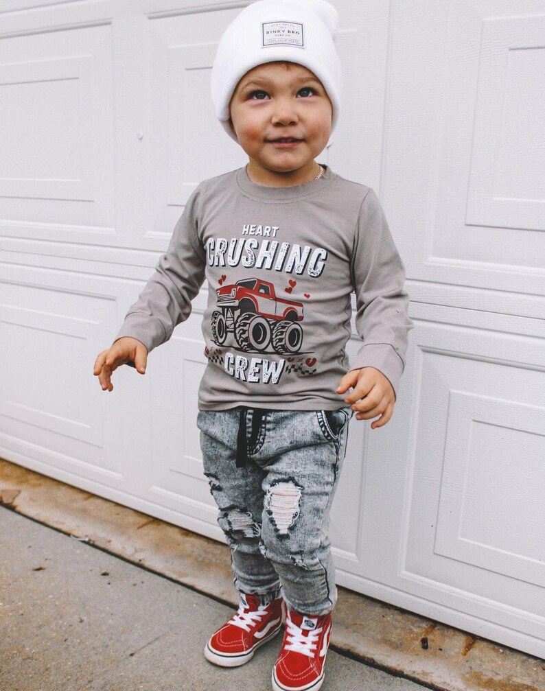 May include: A young child wearing a gray long-sleeve shirt with a red monster truck graphic and the text "Heart Crushing Crew". The child is also wearing a white beanie with a logo, distressed denim jeans, and red sneakers.