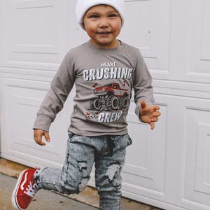 May include: A young child wearing a grey long-sleeve shirt with a monster truck graphic and the text "Heart Crushing Crew". The child is also wearing distressed denim jeans, a white beanie with a small logo, and red sneakers.