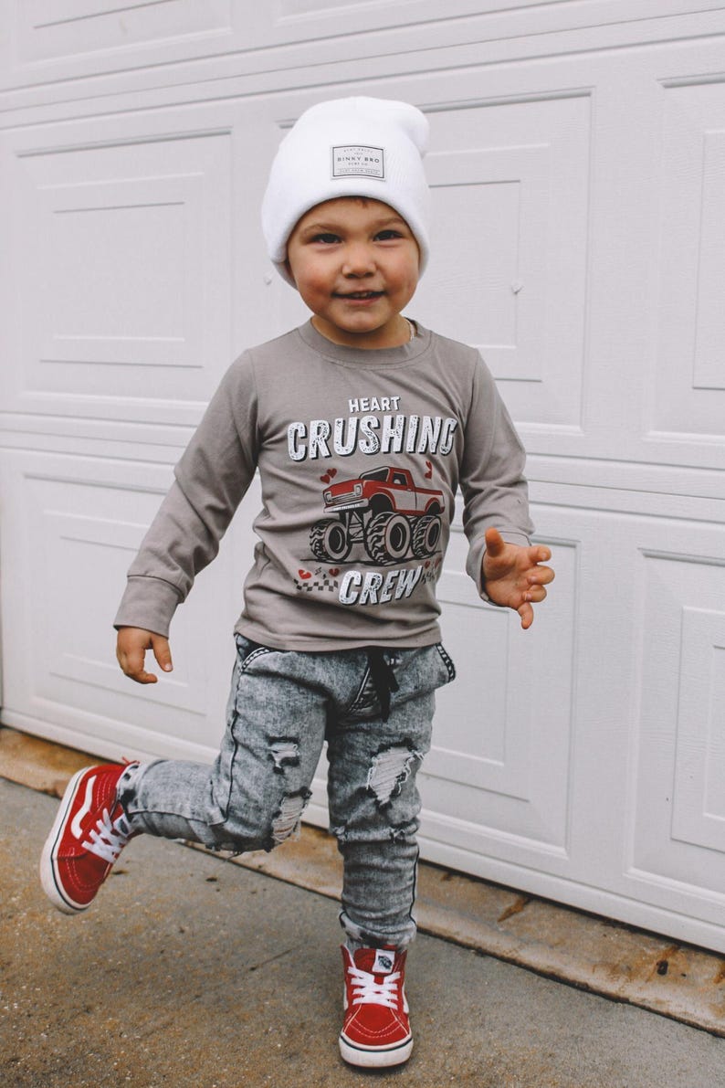 May include: A young child wearing a grey long-sleeve shirt with a monster truck graphic and the text "Heart Crushing Crew". The child is also wearing distressed denim jeans, a white beanie with a small logo, and red sneakers.
