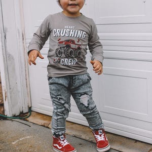 May include: A young child wearing a gray long-sleeve shirt with a red monster truck graphic and the text "Heart Crushing Crew". The child is also wearing blue ripped jeans, a white beanie, and red high-top sneakers.