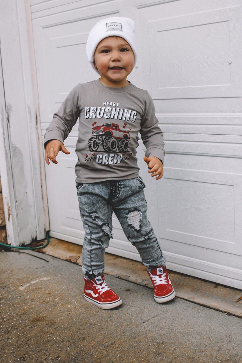 May include: A young child wearing a gray long-sleeve shirt with a red monster truck graphic and the text "Heart Crushing Crew". The child is also wearing blue ripped jeans, a white beanie, and red high-top sneakers.