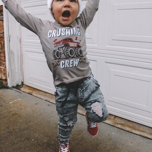 May include: A young child wearing a grey long-sleeved shirt with a red monster truck graphic and the text "The Crushing Crew". The child is also wearing a white beanie, ripped denim jeans, and red high-top sneakers.