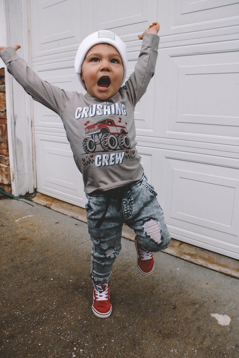 May include: A young child wearing a grey long-sleeved shirt with a red monster truck graphic and the text "The Crushing Crew". The child is also wearing a white beanie, ripped denim jeans, and red high-top sneakers.