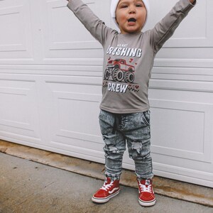 May include: A young child wearing a grey long-sleeved shirt with a red monster truck graphic and the text "Heart Crushing Crew". The child is also wearing a white beanie, distressed denim jeans, and red high-top sneakers.