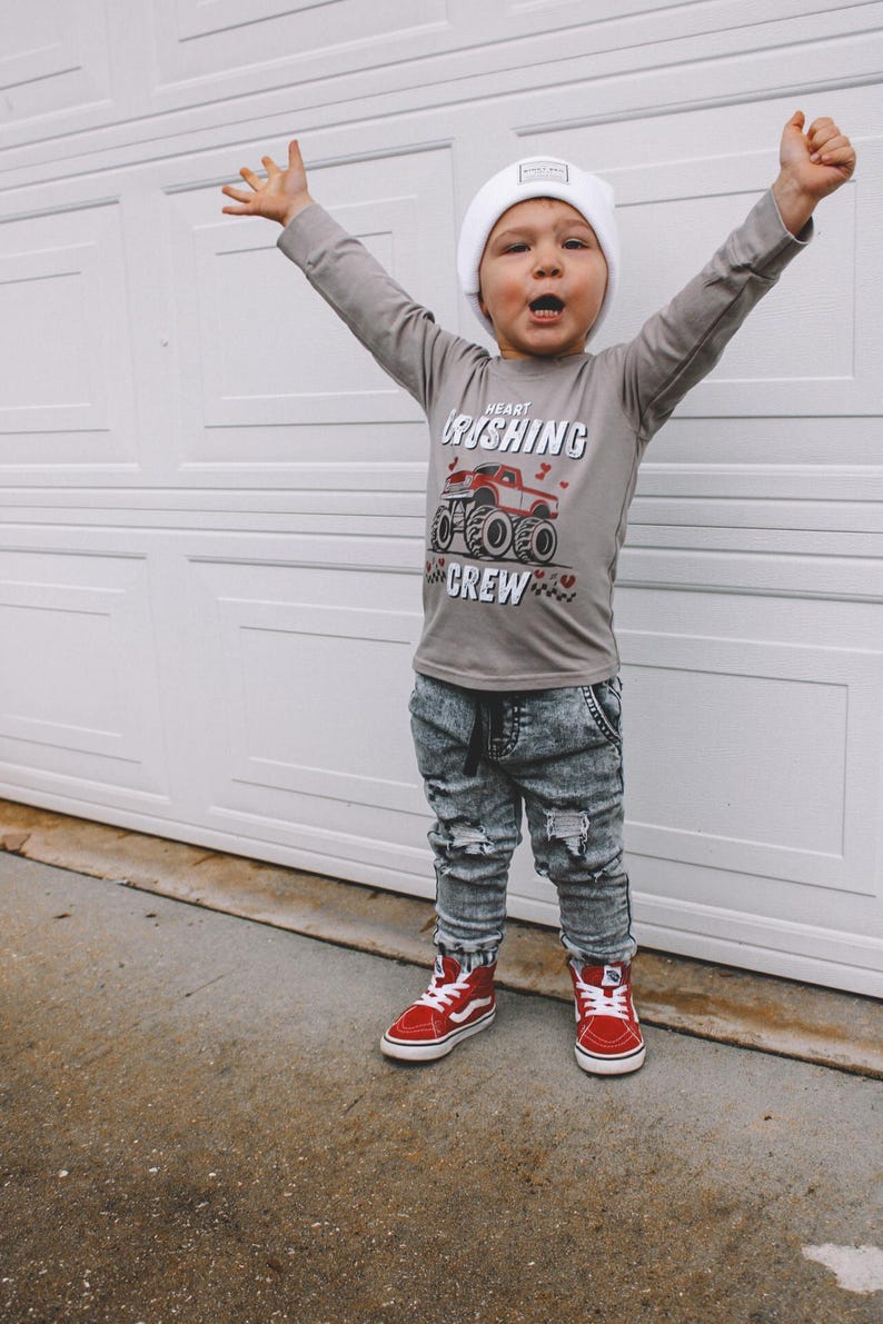 May include: A young child wearing a grey long-sleeved shirt with a red monster truck graphic and the text "Heart Crushing Crew". The child is also wearing a white beanie, distressed denim jeans, and red high-top sneakers.