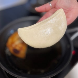 May include: A hand holds up a light brown, crescent-shaped pancake over a black frying pan. The pancake is slightly puffed and has a shiny surface. A smaller, darker pancake is visible in the pan.