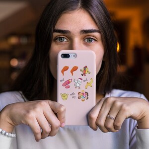 May include: A pink phone case with a variety of colorful stickers, including a chicken, a hot dog, a banana, a hand with a thumbs up, and a peace sign. The text "and my" is visible on the case.
