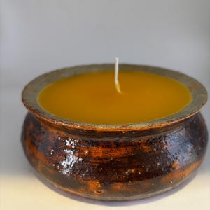 May include: A handmade candle in a rustic, ceramic bowl. The candle wax is a warm yellow-orange color, and the bowl has a dark brown and orange glazed finish. A white wick is centered in the wax.