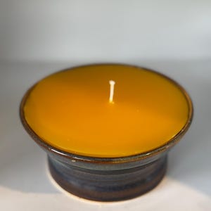 May include: A handmade candle in a ceramic container. The candle has a bright yellow wax and a white wick. The container is a dark blue and brown color. The candle is a decorative item.