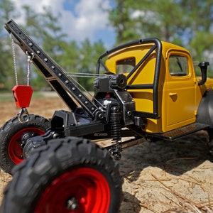 May include: A yellow toy truck with a black roll cage and a black crane arm. The truck has red wheels and is sitting on a bed of pine needles.