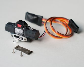 Op de afbeelding: Een zwarte en zilveren elektrische lier met een rode knop en een spoel met wit touw. De lier is verbonden met een set van drie draden met oranje, bruin en gele isolatie.