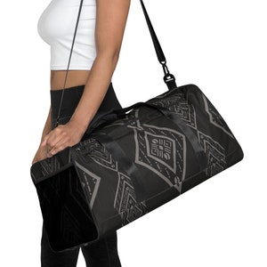 Könnte beinhalten: Eine schwarze und graue Reisetasche mit geometrischem Muster und einem schwarzen Schultergurt. Die Tasche hat einen Reißverschluss und eine große Tasche auf der Vorderseite.