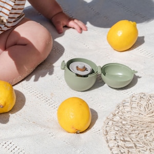 May include: A green plastic baby bottle with a lid and a white background. There are three yellow lemons and a white crocheted bag on the background.