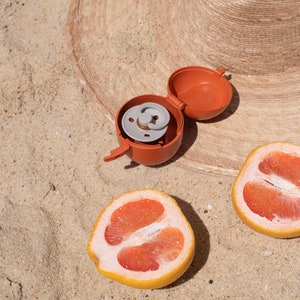May include: A close-up of a red and gray pacifier holder on a sandy beach with two halves of a grapefruit.