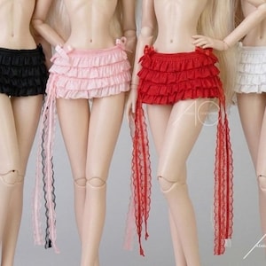 Puede incluir: Cuatro faldas de muñeca en negro, rosa, rojo y blanco. Cada falda presenta múltiples capas de volantes y largas cintas decorativas. Las faldas están atadas con lazos en la cintura. El texto "4C AZJAD COUTURER" es visible en la parte inferior derecha.