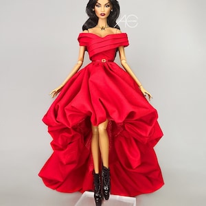 May include: A fashion doll wearing a red off-the-shoulder gown with a high-low hemline and a jeweled crown. The doll is standing on a clear acrylic base.