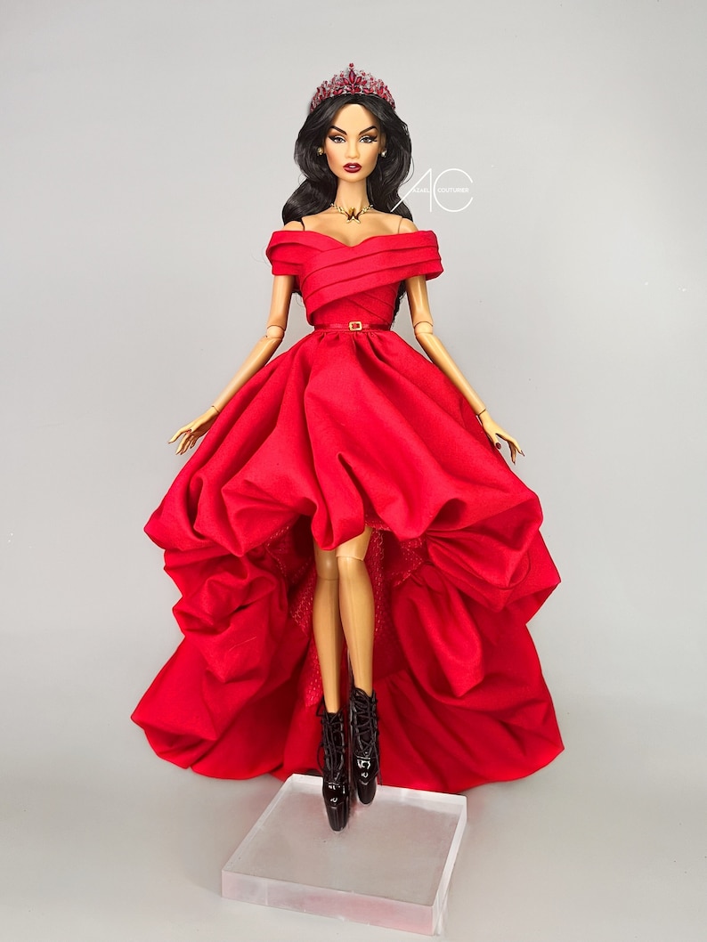 May include: A fashion doll wearing a red off-the-shoulder gown with a high-low hemline and a jeweled crown. The doll is standing on a clear acrylic base.
