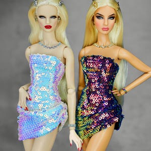 Puede incluir: Dos muñecas de moda con cabello rubio y maquillaje glamuroso. Visten minivestidos sin tirantes con lentejuelas en tonos azules y morados. Cada muñeca tiene joyas, incluyendo collares y accesorios para el cabello. Las muñecas están sobre un fondo gris.