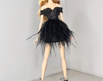 Puede incluir: Una muñeca de moda con un vestido negro de lentejuelas con ribete de plumas y una corona negra. La muñeca tiene el pelo rubio y lleva zapatos negros de tacón.