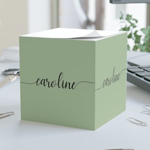 May include: A square, sage green sticky note cube with the name "caroline" in black script on two sides. The cube sits on a white surface with scattered silver paper clips.