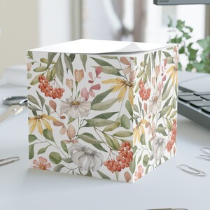 May include: A white cube-shaped notepad with a floral pattern featuring white, yellow, and pink flowers, green leaves, and red berries. The notepad is sitting on a white desk with paper clips and a laptop in the background.