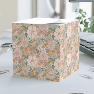 May include: A floral patterned sticky note cube featuring a repeating design of pastel flowers in shades of pink, yellow, and green on a beige background. The cube sits on a white surface with office supplies nearby.