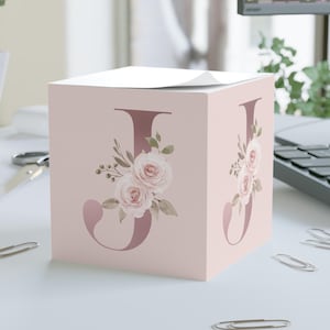 May include: A pink memo cube featuring the letter "J" in a darker pink hue, adorned with a floral design of roses and leaves. The cube sits on a white surface with scattered paper clips, near a laptop and a plant.