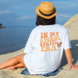 Peut inclure: Un t-shirt blanc avec le texte "IN MY APEROL SPRITZ ERA" en orange et p&ecirc;che. Un verre d'Aperol Spritz avec une tranche d'orange est repr&eacute;sent&eacute; &agrave; c&ocirc;t&eacute; du texte.