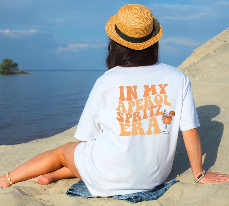 Peut inclure: Un t-shirt blanc avec le texte "IN MY APEROL SPRITZ ERA" en orange et p&ecirc;che. Un verre d'Aperol Spritz avec une tranche d'orange est repr&eacute;sent&eacute; &agrave; c&ocirc;t&eacute; du texte.
