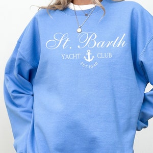 May include: A light blue sweatshirt with the text "St. Barth Yacht Club Est. 1648" in white. The text is in a cursive font and features an anchor symbol.