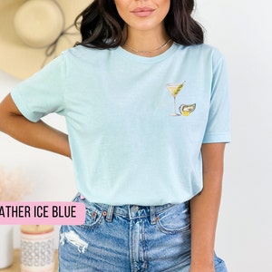 Puede incluir: Una camiseta azul claro con un gr&aacute;fico de una copa de martini y una ostra. El texto "HEATHER ICE BLUE" es visible en la parte inferior de la imagen.