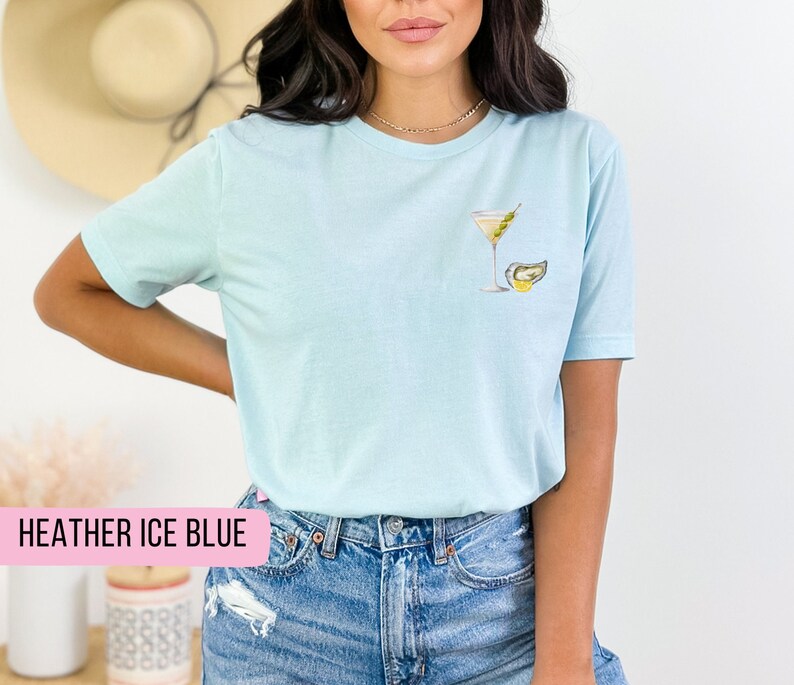 Puede incluir: Una camiseta azul claro con un gr&aacute;fico de una copa de martini y una ostra. El texto "HEATHER ICE BLUE" es visible en la parte inferior de la imagen.