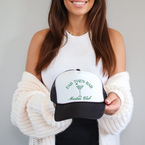 Op de afbeelding: Een zwart-witte trucker-pet met een groen logo met de tekst "Par Then Bar Martini Club" en een groene martini-glasafbeelding.