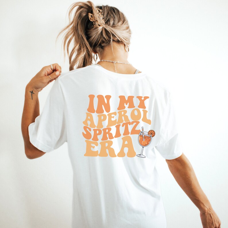 Peut inclure: T-shirt blanc avec le texte "IN MY APEROL SPRITZ ERA" dans une police r&eacute;tro avec un sch&eacute;ma de couleurs orange et jaune. Un verre d'Aperol Spritz est repr&eacute;sent&eacute; &agrave; c&ocirc;t&eacute; du texte.