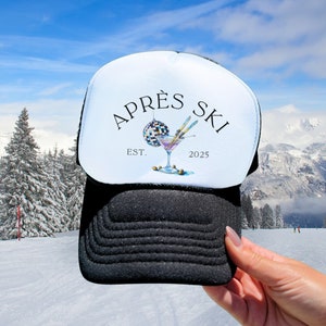 May include: White trucker hat with black mesh back. The front of the hat features a black and white illustration of a martini glass with a disco ball and skis. The text "APRÉS SKI" is above the illustration and "EST. 2025" is below.
