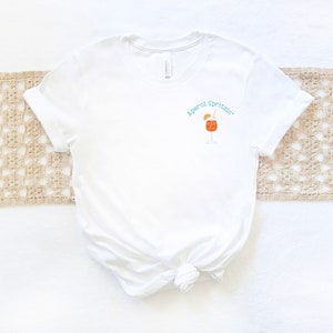 May include: White t-shirt with a graphic of an Aperol Spritz cocktail and the text "Aperol Spritzin'" in turquoise.