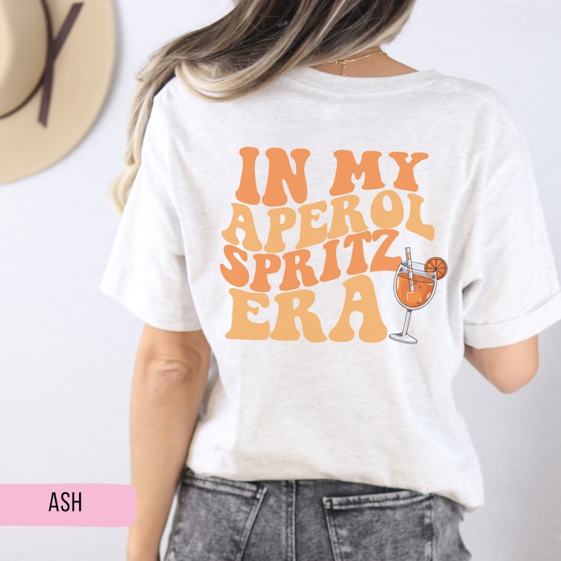 Peut inclure: Un t-shirt blanc avec le texte "IN MY APEROL SPRITZ ERA" en lettres orange et jaunes. Un verre d'Aperol Spritz avec une tranche d'orange et une paille est &agrave; droite du texte.