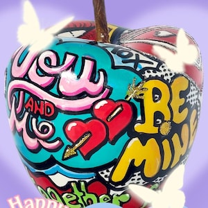 Könnte beinhalten: Ein roter, gelber, blauer und grüner Apfel mit den Worten "You and Me" und "Be Mine" darauf. Der Apfel ist mit Herzen, Pfeilen und anderen Valentinstags-Grafiken verziert. Der Hintergrund ist helllila mit den Worten "Happy Valentine's Day" in rosa.