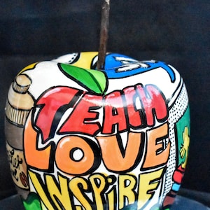 Può includere: Una mela di ceramica dipinta con parole colorate, tra cui "Teach", "Love" e "Inspire". La mela ha un gambo verde ed è decorata con una varietà di motivi.