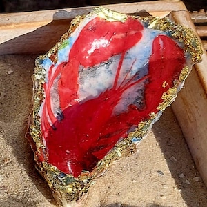 Könnte beinhalten: Ein roter Hummer, der auf eine weiße Austernschalen mit goldenen Blattverzierungen gemalt ist. Die Schale liegt auf einer sandigen Oberfläche.