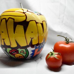May include: A yellow apple with a black and red graphic design that says "AKAW" on it. The apple is next to two red tomatoes.