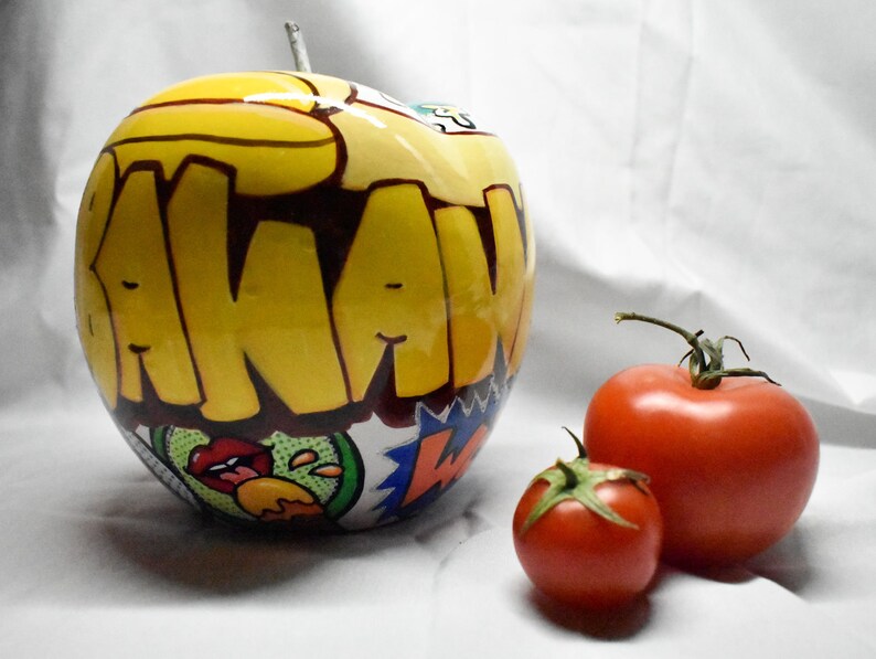 May include: A yellow apple with a black and red graphic design that says "AKAW" on it. The apple is next to two red tomatoes.
