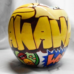 May include: A yellow ceramic apple with the word "AWAW" written in brown letters on the front. The apple has a colorful comic book design with a green, yellow, and blue pattern.