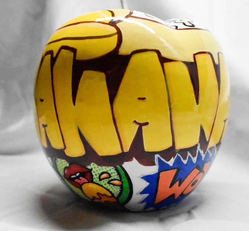 May include: A yellow ceramic apple with the word "AWAW" written in brown letters on the front. The apple has a colorful comic book design with a green, yellow, and blue pattern.
