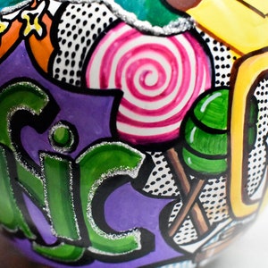 May include: A purple, green, yellow, and white painted ornament with a swirl design, polka dots, and the word "UNIC" in green letters.