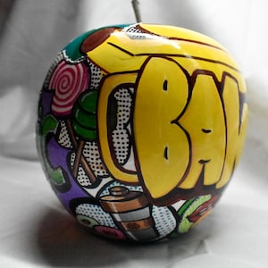 May include: A yellow apple with a colorful pop art design. The apple is painted with a variety of images, including a candy cane, a lollipop, and the word "BAW" in graffiti style.