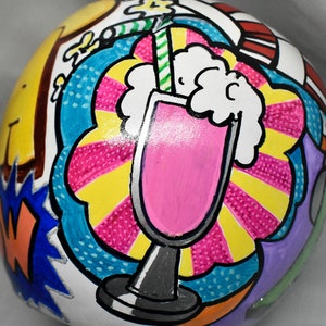 May include: A white ceramic ball with a hand-painted design of a pink milkshake with whipped cream and a straw. The background is a colorful comic book style pattern with yellow, blue, pink, and red.
