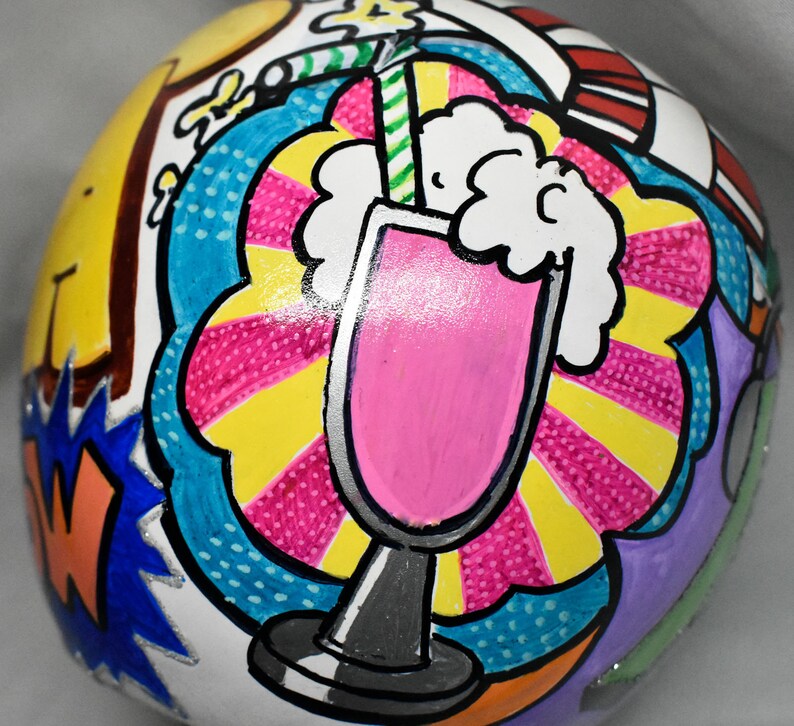 May include: A white ceramic ball with a hand-painted design of a pink milkshake with whipped cream and a straw. The background is a colorful comic book style pattern with yellow, blue, pink, and red.